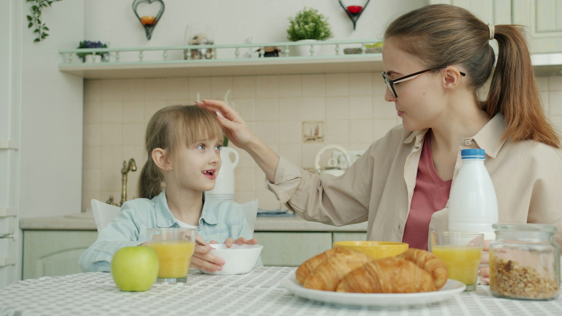 A parent smoothing her daughter's hair across the breakfast table at home